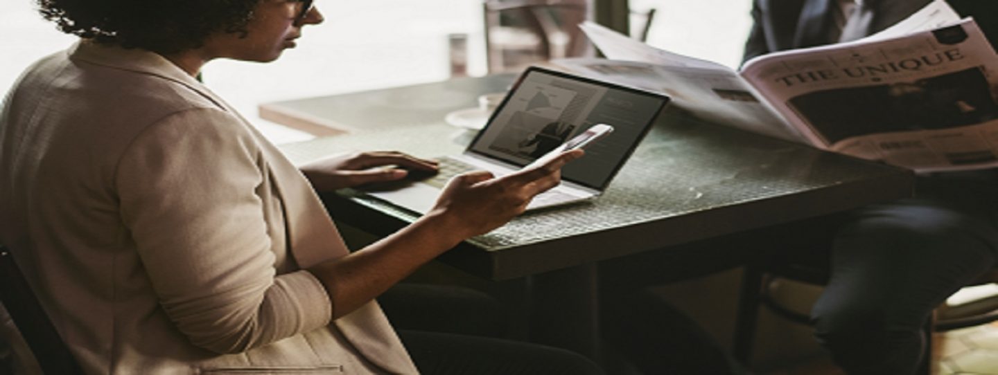 a woman in front of an open laptop using her mobile phone and a man opposite her reading newspaper