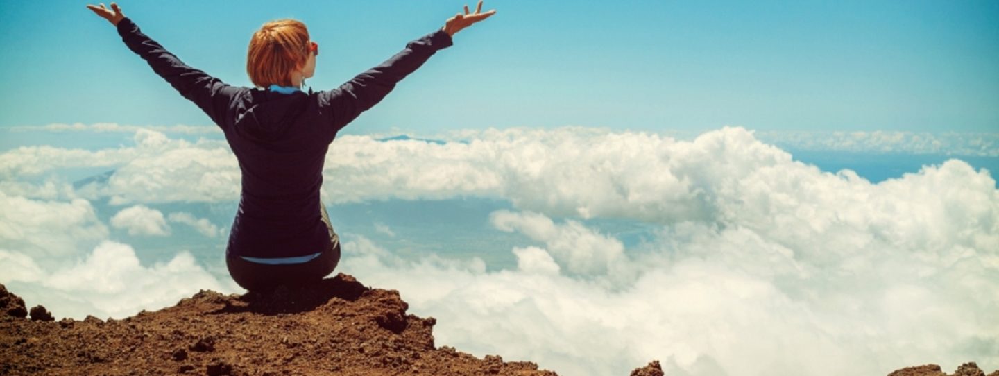 woman sitting on cliff raising both hands towards sky with clouds