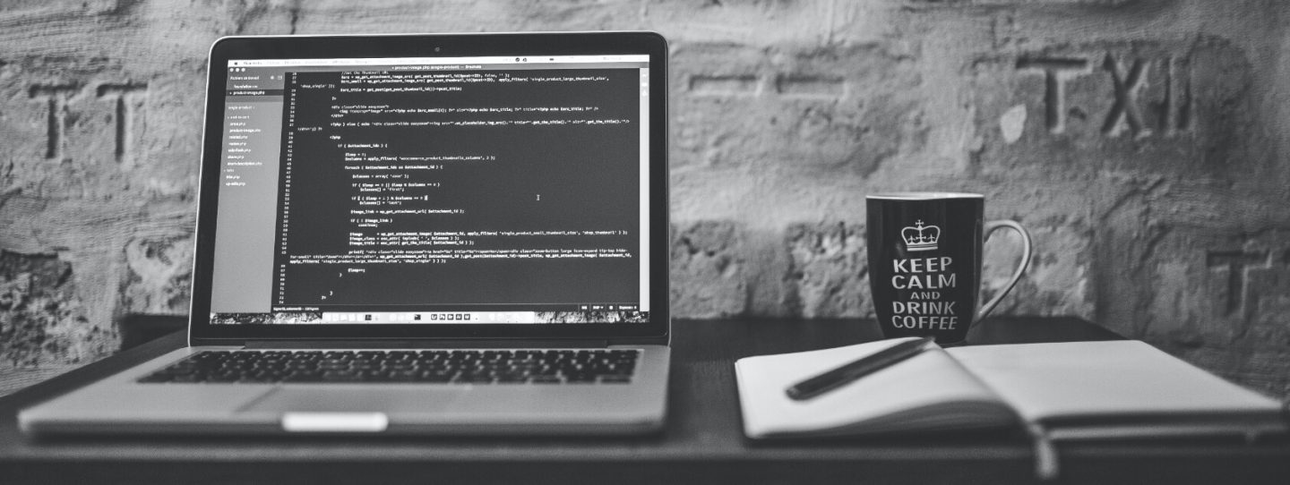 a laptop with code lines on the screen and a notebook, pen and coffee mug set on a work station